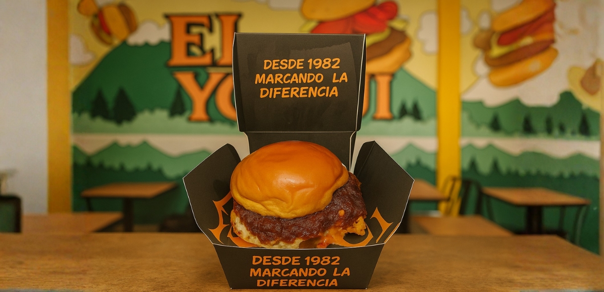 Hamburguesa en pan brioche dentro de caja negra abierta con salsa y queso; fondo del local con mural desenfocado.