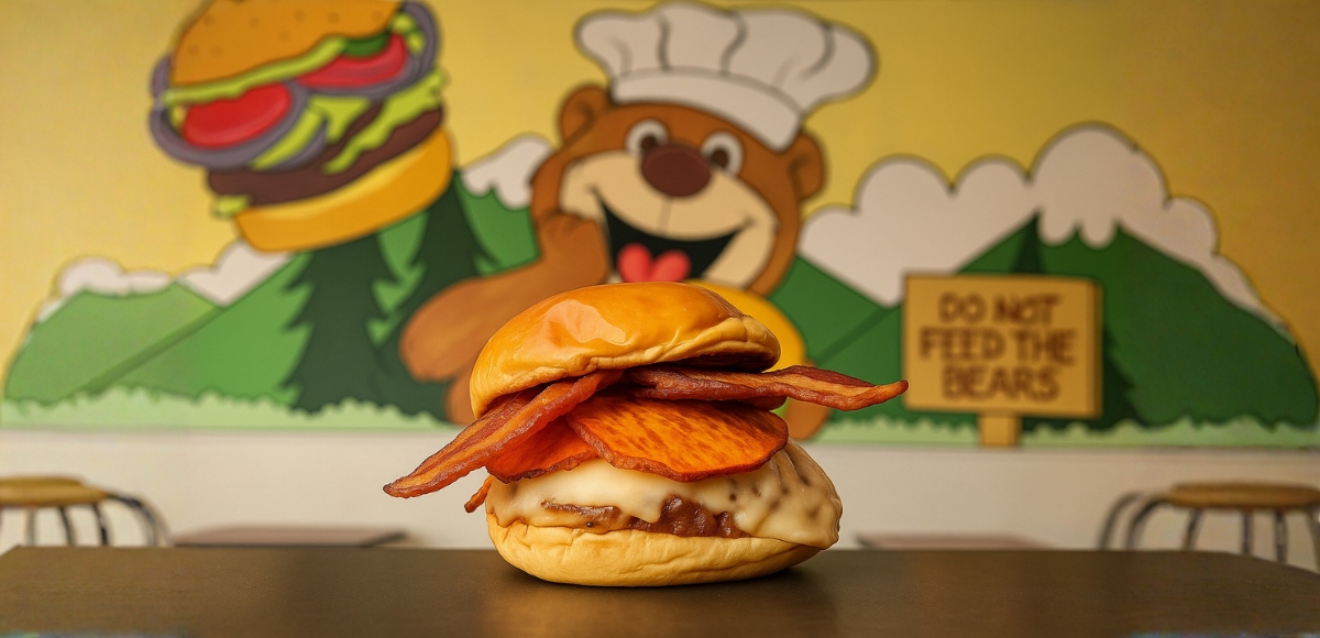Hamburguesa con bacon crujiente, lámina de boniato y queso fundido en pan brioche, con mural de oso desenfocado al fondo.