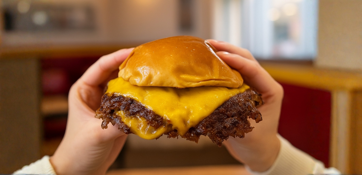 Manos sosteniendo una hamburguesa con queso cheddar fundido y bordes crujientes de carne en pan brioche, primer plano en interior.