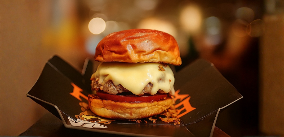 Hamburguesa con queso fundido y rodaja de tomate en pan brioche, presentada en una caja negra, primer plano en interior.